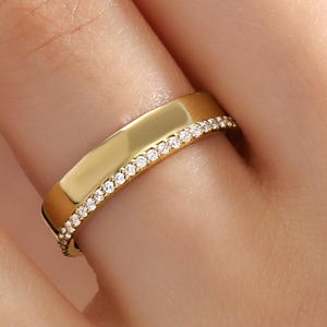 May include: A gold-colored ring with a flat top and a band of small, clear gemstones. The ring is on a finger, showcasing its design and sparkle. The ring's band is approximately 0.25 inches wide.