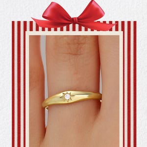 May include: A gold ring with a starburst design and a small diamond. The ring is on a finger, set against a red and white striped background with a red bow. The words "Merry Christmas" are written in red.