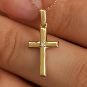 May include: A gold cross pendant suspended from a delicate gold chain. The cross features a smooth, polished finish with a single small, clear, round-cut gemstone set at the center where the vertical and horizontal bars intersect.