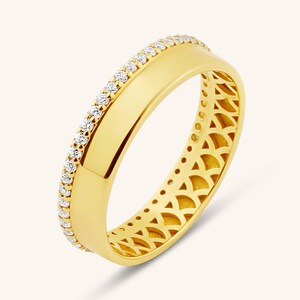 May include: A yellow gold band ring featuring a single row of small, round, clear pavé-set stones along one edge. The band has a smooth, polished surface on one side and a decorative, open-work lattice pattern along the inner and outer edges.