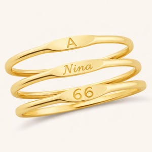 May include: Three gold-colored rings stacked together. Each ring has an oval-shaped design with engraved text. The top ring has the letter "A", the middle ring has the name "Nina", and the bottom ring has the number "66".