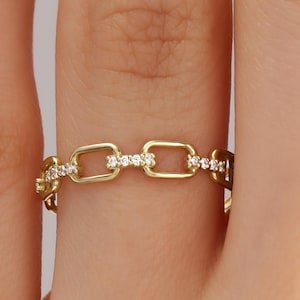 May include: A gold chain link ring with small, clear gemstones set into the links. The ring is shown on a finger, with the chain link design wrapping around the finger. The gemstones add sparkle to the ring.