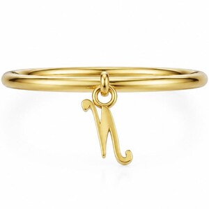 May include: A polished gold ring featuring a thin, smooth band and a single dangling charm in the shape of the letter N. The charm is attached to the band by a small circular jump ring, creating a delicate and minimalist piece of jewellery.