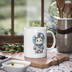 May include: White ceramic mug with a cartoon illustration of a rabbit wearing an astronaut suit and helmet. The rabbit is white with pink ears and nose. The astronaut suit is blue and white with orange accents. The background is white.