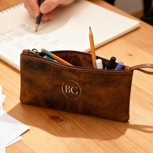 Puede incluir: Un estuche de lápices de cuero marrón con cremallera, que contiene lápices, bolígrafos y otros artículos de papelería. El estuche tiene un emblema circular dorado con las letras "BG".