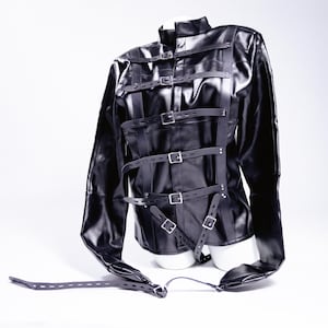 Puede incluir: Una chaqueta negra de manga larga con múltiples correas de cuero y hebillas plateadas. La chaqueta se exhibe en un maniquí blanco y tiene un cuello alto. Las mangas se extienden a los lados, con correas colgando.