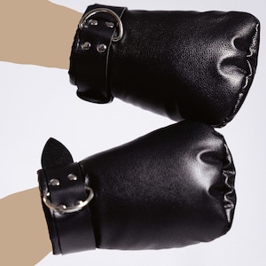 Puede incluir: Un par de esposas de cuero negro. Cada esposa tiene una correa ancha con una hebilla de metal y un anillo en D. Los puños acolchados están fruncidos en el extremo. Las esposas están diseñadas para usarse en las muñecas.