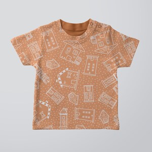 Op de afbeelding: Een bruin T-shirt met korte mouwen met een wit peperkoekhuispatroon. De huizen zijn wit omlijnd en hebben kleine witte stippen die over het hele ontwerp zijn verspreid.