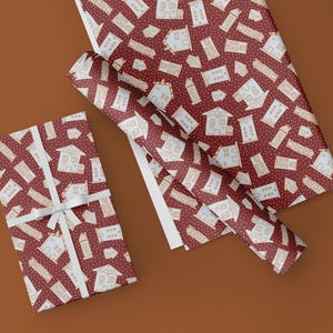 Op de afbeelding: Twee rollen rood inpakpapier met een wit patroon van huizen en stippen. Het papier is gevouwen en wordt getoond op een bruin oppervlak.