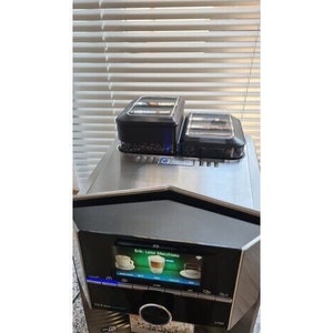 May include: A black and silver espresso machine with a digital display showing the text "Eink Latte Macchiato". The machine has two black containers on top.