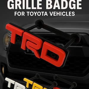 May include: A black plastic TRD grille badge for Toyota vehicles. The badge is red, yellow, and white. The badge is mounted on a black truck grille.