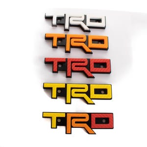 Peut inclure: Collection de cinq emblèmes TRD de différentes couleurs : blanc, orange, rouge, jaune et un dégradé jaune-orange-rouge. Chaque emblème a une base noire avec des trous de montage. Le logo TRD est bien visible.