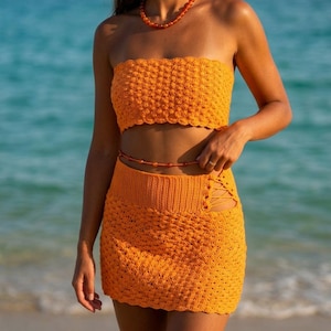 Peut inclure: Un ensemble deux pièces en crochet orange vif composé d'un haut bandeau sans bretelles et d'une mini-jupe assortie. La jupe présente une large ceinture côtelée et une découpe latérale avec des lacets croisés. Le tissu a un motif texturé.