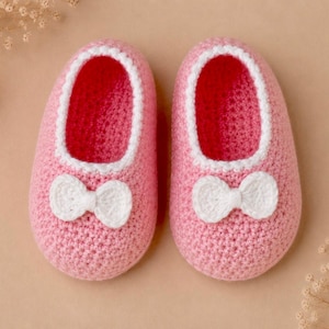 May include: A pair of handmade crochet baby booties in a soft pink yarn. Each shoe features a white crocheted trim around the opening and a small, white crocheted bow centered on the front toe area. The texture is consistent and uniform.