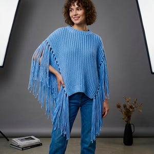 Op de afbeelding: Een lichtblauwe gebreide trui met franjedetails. De trui heeft een ronde halslijn en lange franjes die aan de mouwen en zijkanten hangen. Het model draagt een blauwe jeans. De achtergrond is effen grijs.