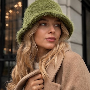 May include: A fuzzy olive green bucket hat with a textured, shaggy yarn finish. The hat features a rounded crown and a downward-sloping brim, crafted from a soft, fibrous material that creates a distinct, tactile appearance across the entire surface.