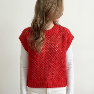 May include: A bright red, sleeveless knit top with a textured, open weave design. The top has a crew neck and a ribbed hem. The model is wearing a long-sleeved white shirt underneath the red knit top.