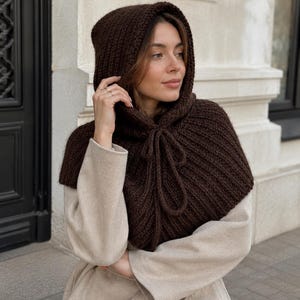 Op de afbeelding: Een donkerbruine gebreide capuchon met een geribbelde textuur en een bijpassend trekkoord bij de hals. Het kledingstuk is ontworpen om het hoofd en de schouders te bedekken en biedt een gelaagde look over een lichtbeige jas met lange mouwen.