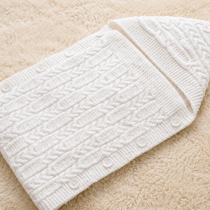 May include: A white knitted baby sleeping bag with a hood and a tassel. The sleeping bag has a cable knit design and is secured with buttons. The item is lying on a cream-colored, fluffy surface.