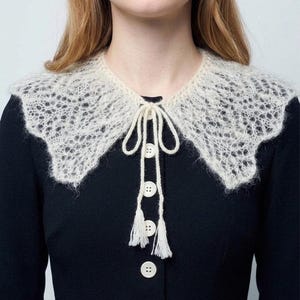 May include: A cream-colored, open-weave collar with a scalloped edge, tied with a delicate string and adorned with three small buttons and tassels. The collar is worn over a dark navy blue garment.