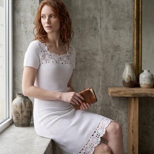 May include: White crochet dress with a square neckline and short sleeves. The dress features a decorative lace trim at the neckline and hem. A woman is holding a brown leather-bound book. The dress is knee-length.