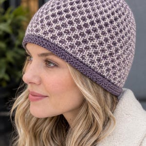 Puede incluir: Un gorro de punto con un patrón de panal texturizado en tonos morado y crema. El gorro tiene un borde acanalado de color morado oscuro sólido que rodea la base, proporcionando un ajuste estructurado. El diseño general es uniforme.