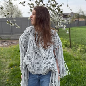 May include: A light gray, chunky knit sweater with long fringe detailing. The sweater is worn over blue jeans. The background features a green lawn, a fence, and flowering tree branches.
