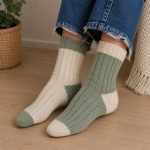 May include: A pair of ribbed knit socks featuring a color-blocked design. One sock is primarily cream with a sage green cuff and toe, while the other is sage green with a cream cuff and toe. The socks are worn with blue denim jeans.
