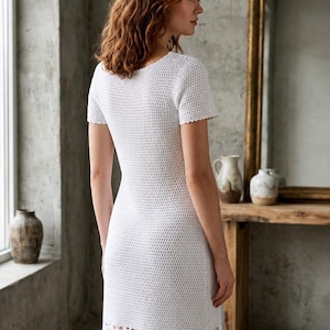 May include: A white, short-sleeved crochet dress with a scalloped hem. The dress has a fitted silhouette and a scoop neckline. The model is standing in front of a window and a wooden table with a mirror.