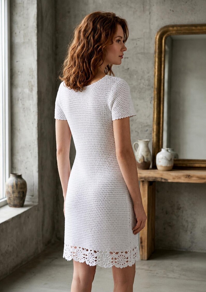 May include: A white, short-sleeved crochet dress with a scalloped hem. The dress has a fitted silhouette and a scoop neckline. The model is standing in front of a window and a wooden table with a mirror.