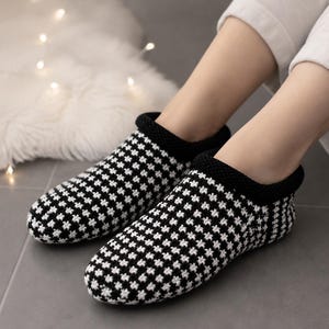 May include: A pair of ankle-high house slippers featuring a black and white houndstooth pattern. The slippers are constructed with a knitted fabric and include a solid black ribbed trim around the opening, providing a snug fit around the ankles.