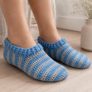 May include: A pair of handmade crochet slippers featuring alternating horizontal stripes of light blue and grey yarn. The slippers have a ribbed cuff around the ankle opening and a rounded toe design, crafted with a textured stitch pattern throughout.