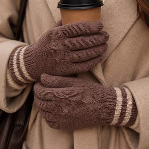 May include: A pair of brown knit gloves featuring a ribbed cuff with two horizontal beige stripes. The gloves are constructed with a standard five-finger design and a consistent knit texture throughout, providing a simple and functional hand covering.
