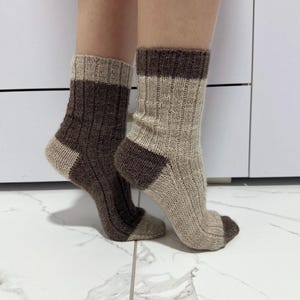 Puede incluir: Un par de calcetines de punto acanalado en tonos marrón y beige. Los calcetines tienen un puño, talón y puntera de color marrón oscuro, con un cuerpo beige más claro. Los calcetines se exhiben sobre un suelo de baldosas blancas.