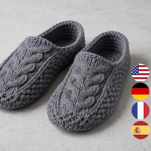 May include: A pair of grey crochet slippers featuring a prominent central cable knit pattern running along the top of the foot, with a textured stitch design on the sides and a rounded toe shape, presented in a neutral grey yarn colour.