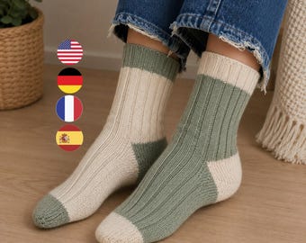 May include: A pair of ribbed knit socks featuring a color-blocked design. One sock is primarily cream with a sage green cuff, heel, and toe, while the other is sage green with a cream cuff, heel, and toe. The socks are worn with blue jeans.