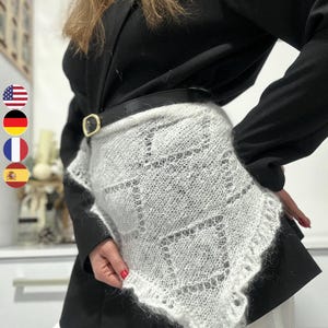 May include: A white, open-weave shawl with a geometric pattern and a fringed edge. The shawl is worn over a black blazer and a black belt with a gold buckle. The outfit is completed with white pants.