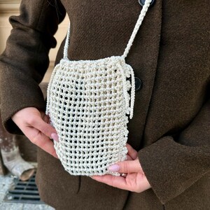 May include: Off-white, open-weave crochet bag with a long strap. The bag is held against a brown coat. The bag's design features a series of evenly spaced holes, creating a net-like appearance. The strap is made of the same material as the bag.