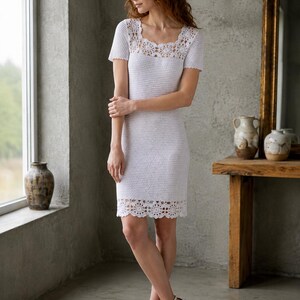 May include: A white crochet dress with a square neckline and short sleeves. The dress features a decorative lace trim at the neckline and hem. The woman is wearing white high-heeled sandals. The dress is knee-length.