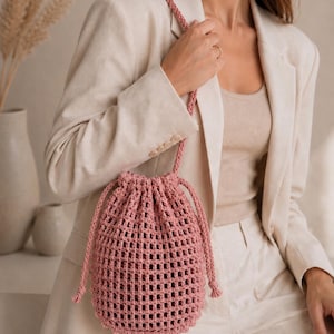 May include: A dusty rose drawstring bucket bag crafted from a thick, open-weave cord material. The bag features a grid-like pattern, a circular base, and a matching braided shoulder strap that cinches the top opening with two knotted ends.