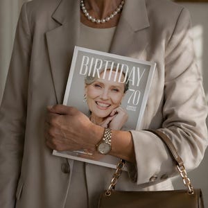 Puede incluir: Una mujer con un traje beige sostiene una revista con el texto "BIRTHAY 70 LIMITED EDITION". Lleva un collar de perlas, un reloj dorado y pendientes de aro. Se ve un bolso marr&oacute;n con una cadena dorada.