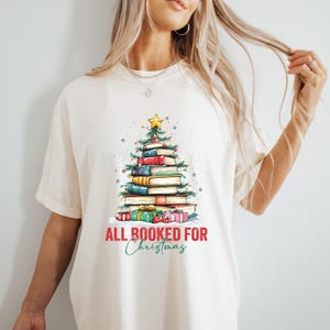 May include: Cream-colored t-shirt featuring a Christmas tree made of stacked books. The tree is topped with a gold star and decorated with lights. The phrase "ALL BOOKED FOR Christmas" is printed below the tree in red and green.