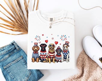 May include: White Comfort Colors t-shirt featuring a graphic of six dogs dressed in patriotic attire. The dogs are wearing hats, bow ties, and sweaters with red, white, and blue designs. Fireworks and heart-shaped balloons are also included in the design.