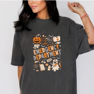 May include: Dark grey t-shirt featuring a Halloween-themed design. The graphic includes a pumpkin, medical symbols, and the words "EMERGENCY DEPARTMENT" in orange and grey. A ghost character is also present.