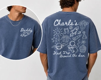 May include: A blue t-shirt with the word "Daddy" and a rocket ship graphic on the front. The back of the shirt features the text "Charls's First Trip Around the Sun" with various space-themed illustrations in white.
