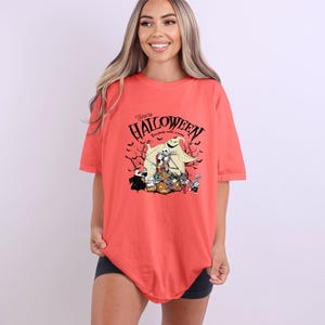Op de afbeelding: Koraalrood T-shirt met een Halloween-thema. Het ontwerp bevat personages en de tekst "Welcome to Halloween, Everybody make a scene". Het shirt heeft korte mouwen en een relaxte pasvorm.