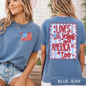 A woman wearing a blue t-shirt with an American flag design and text on the back, standing in front of a brick wall with an American flag visible.