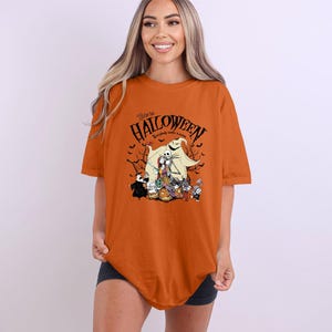 Op de afbeelding: Een oranje t-shirt met een Halloween-thema. Het ontwerp bevat personages en de tekst "Welcome to Halloween, everybody makes a scene". Het shirt heeft een losse pasvorm.