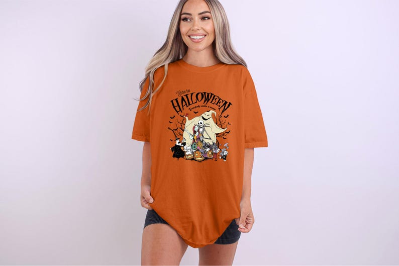Op de afbeelding: Een oranje t-shirt met een Halloween-thema. Het ontwerp bevat personages en de tekst "Welcome to Halloween, everybody makes a scene". Het shirt heeft een losse pasvorm.