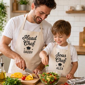 May include: A father and child wearing matching beige aprons in a kitchen. The father's apron reads "Head Chef Daddy" and the child's reads "Sous Chef Alex." They are preparing a salad with fresh vegetables. A glass bowl of salad is on the wooden counter.